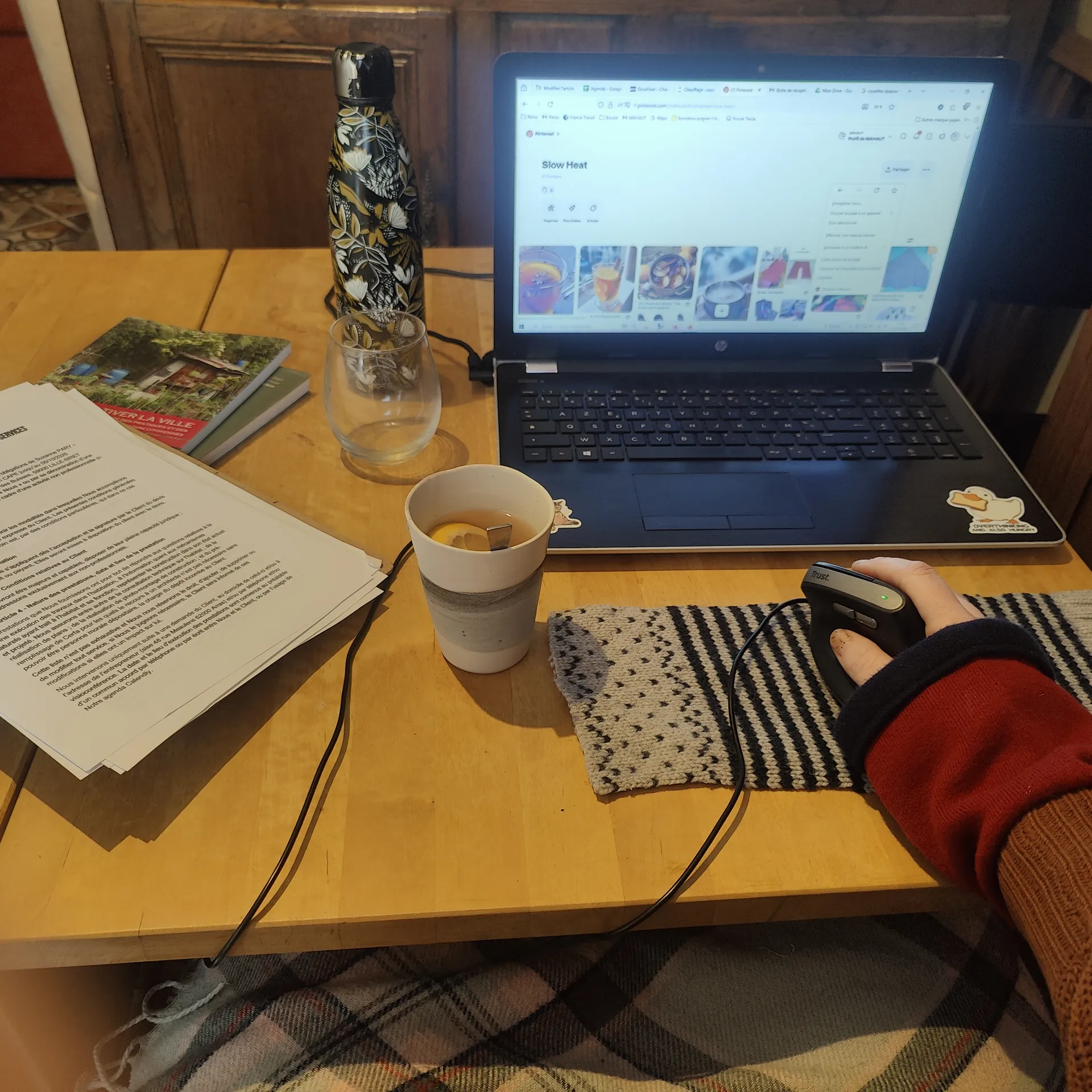 Poste de travaille de bureau. Avec une boisson chaude, un plaid sur les jambes, la main tenant la souris a des sous-manches sous son pull en tricot, le tapis de souris et un rectangle de laine. L'ordinateur est allumé à droite et une pile de documents se trouve à gauche.