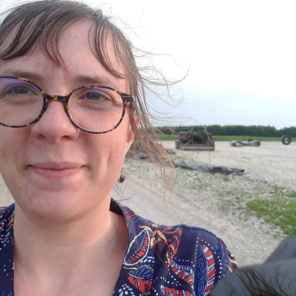 On voit une femme d'une bonne trentaine d'année avec des lunettes écailles, une frange et quelques cheveux bruns qui volent et une boucle d'oreille tombante derrière laquelle on voit une plage et un ciel blanc.