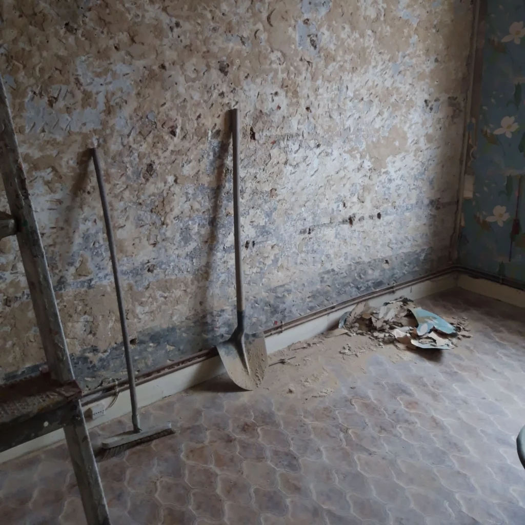 Sol en vinyle, mur en torchis détapissé. Un tas de poussières et papier est visible dans le fond, une pelle et un balai sont posés contre le mur, on aperçoit un escabeau au 1ᵉʳ plan.