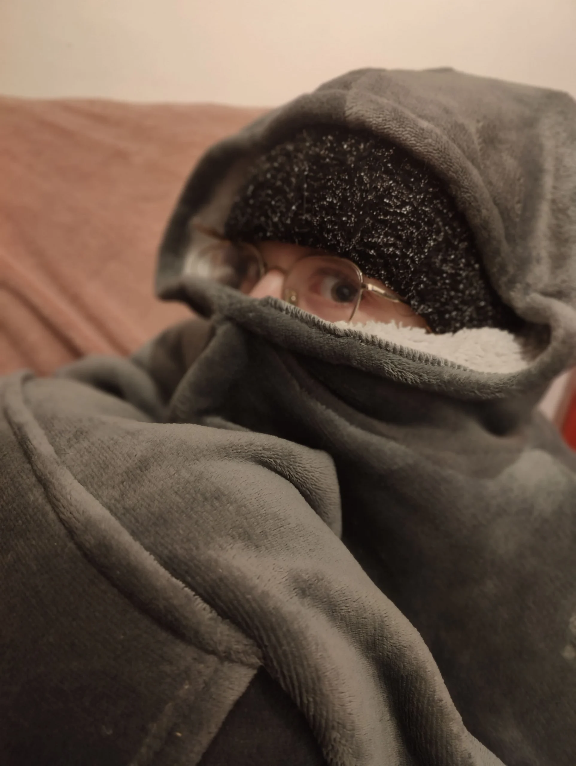 On voit une fille en boule dans un pull en moumoute avec la capuche relevé sur un bonnet tricoté et remonté sur son nez. Seuls les yeux et lunettes paraissent.