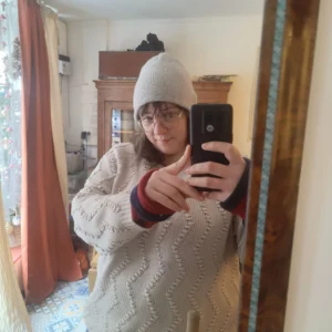 Portrait pris à l'aide d'un miroir sur lequel on voit une femme tenir son portable avec un pull et un bonnet en tricot.