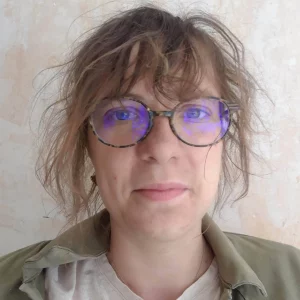 Visage d'une femme d'une trentaine d'années avec des lunettes le visage plein de poussière et quelques cheveux bruns volants en bataille.