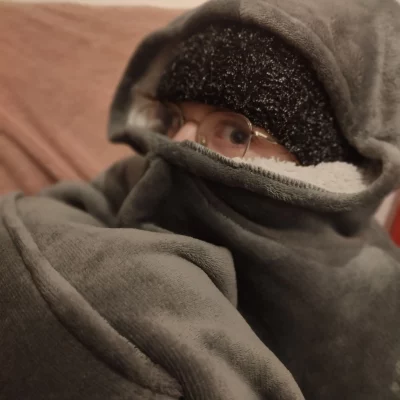 On voit une fille en boule dans un pull en moumoute avec la capuche relevé sur un bonnet tricoté et remonté sur son nez. Seuls les yeux et lunettes paraissent.